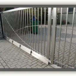 Przesuwna brama wjazdowa ze stali nierdzewnej z pionowymi prętami i łukowatym wzorem, widoczna na tle kostki brukowej i zieleni.