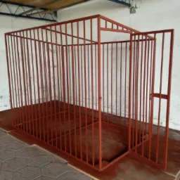 Metalowa konstrukcja klatki pomalowana na czerwono, z otwartymi drzwiami, umieszczona na drewnianej podłodze w pomieszczeniu z betonową posadzką i białą ścianą.