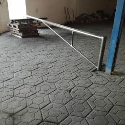 D1 - Surowa stalowa barierka ochronna w hali przemysłowej, widoczne palety i narzędzia w tle, posadzka z betonowej kostki brukowej.