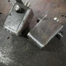 D1 - Dwa surowe, spawane, metalowe prostopadłościany z wystającymi elementami cylindrycznymi leżące na metalowej, chropowatej powierzchni roboczej.