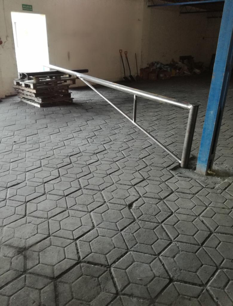 Surowa stalowa barierka ochronna w hali przemysłowej, widoczne palety i narzędzia w tle, posadzka z betonowej kostki brukowej.