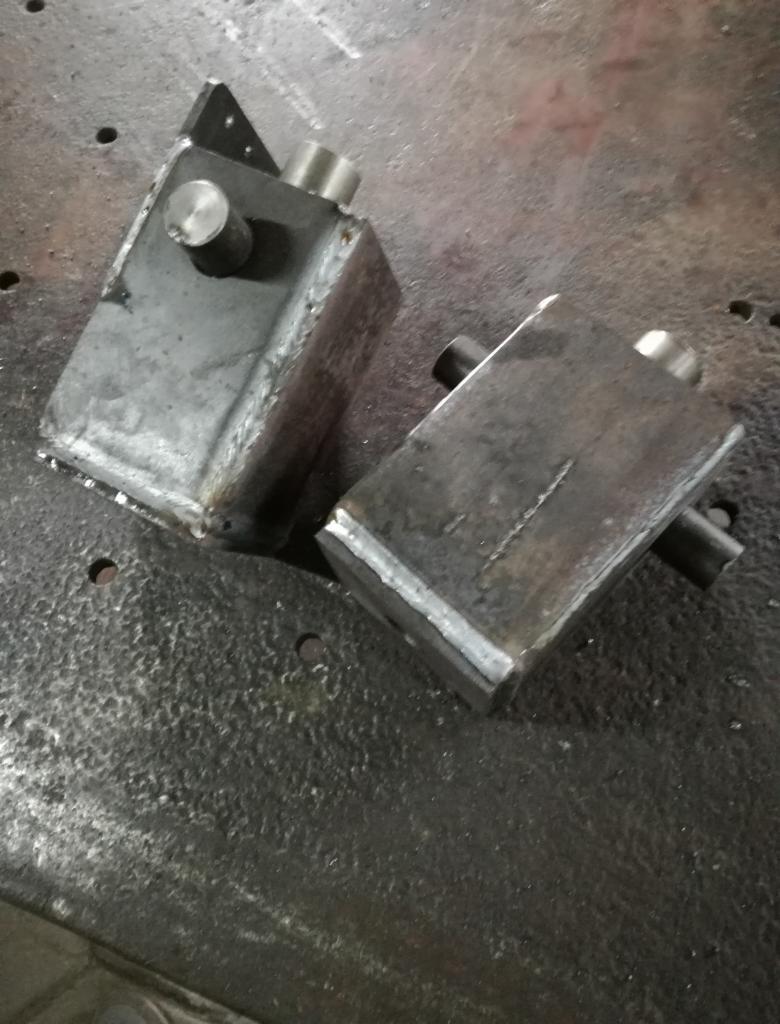 Dwa surowe, spawane, metalowe prostopadłościany z wystającymi elementami cylindrycznymi leżące na metalowej, chropowatej powierzchni roboczej.