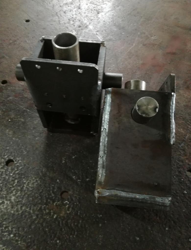 Dwa spawane elementy metalowe, jeden z rurą wewnątrz, leżące na metalowej powierzchni z widocznymi śladami obróbki.