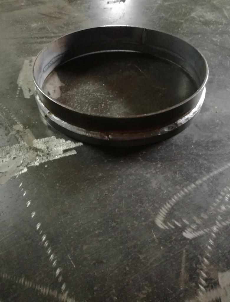 Metalowy pierścień spawany z dwóch elementów na brudnym, metalowym stole warsztatowym, widoczne ślady spawania.