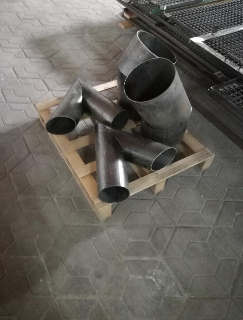 Surowe, metalowe rury i elementy spawane, leżące na drewnianej palecie w warsztacie, z widocznymi śladami obróbki i niedoskonałościami spawalniczymi.