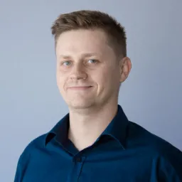 Portret uśmiechniętego mężczyzny w niebieskiej koszuli na tle jasnoszarej ściany.