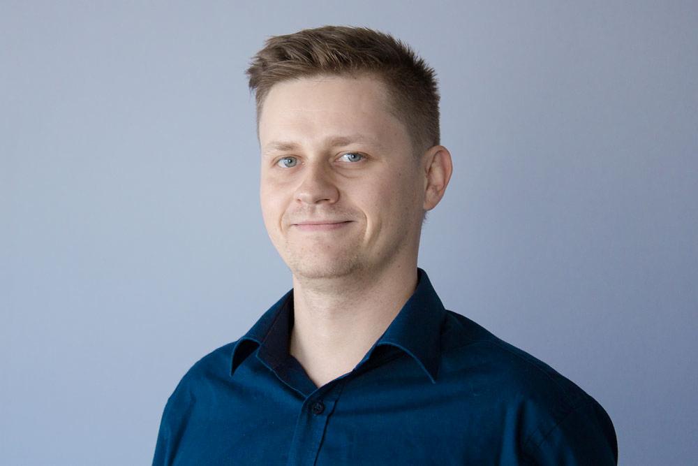 Portret uśmiechniętego mężczyzny w niebieskiej koszuli na tle jasnoszarej ściany.