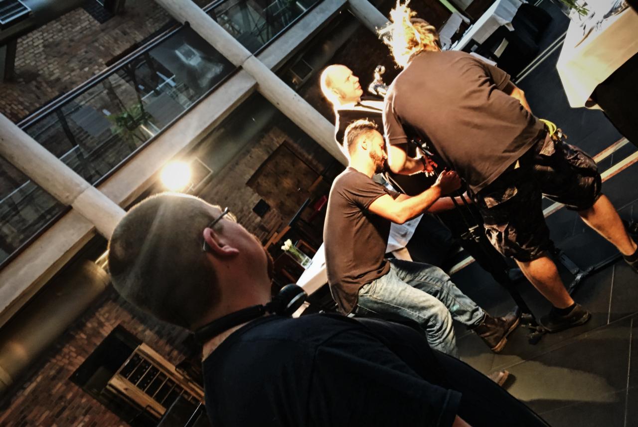 Ekipa filmowa w trakcie pracy: operator kamery z dreadami, dźwiękowiec z mikrofonem, oświetleniowiec i reżyser w nietypowym wnętrzu z ceglanymi ścianami i szklanym sufitem.