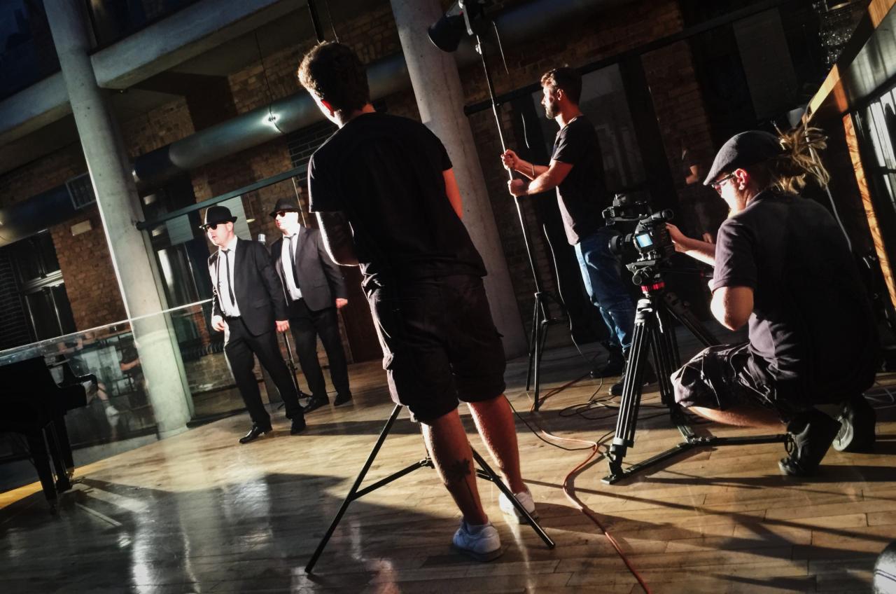 Ekipa filmowa kręci scenę z trzema mężczyznami w czarnych garniturach i kapeluszach typu fedora, przypominających The Blues Brothers, na tle industrialnego wnętrza z ceglanymi ścianami i drewnianą...