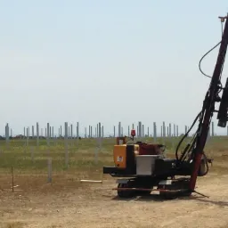 Maszyna wbijająca pale pod konstrukcję, prawdopodobnie pod panele słoneczne, widoczne liczne wbite słupy w tle, teren nierówny, trawiasty.