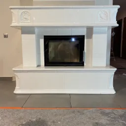 Biała obudowa wkładu kominkowego z ceramicznych płytek, zdobiona reliefami z motywem ptaków, przed położeniem podłogi.