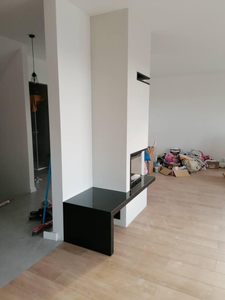 Nowoczesny wkład kominkowy z czarnym blatem w minimalistycznym salonie podczas remontu, widoczne narzędzia i materiały budowlane.