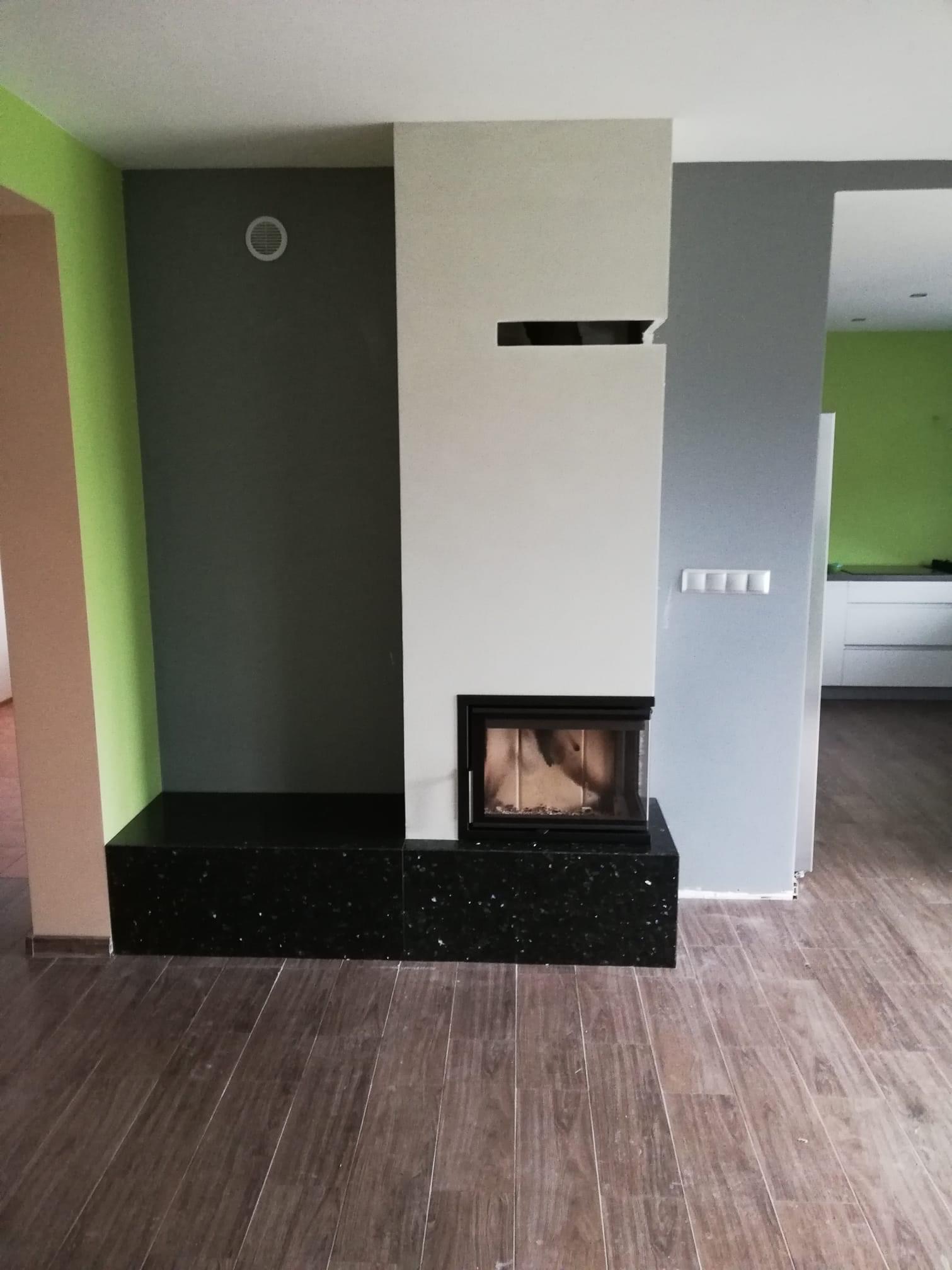 Nowoczesny kominek obudowany czarnym, błyszczącym kamieniem, z częściowo widocznym paleniskiem, w pomieszczeniu z zieloną i szarą ścianą oraz panelami podłogowymi.