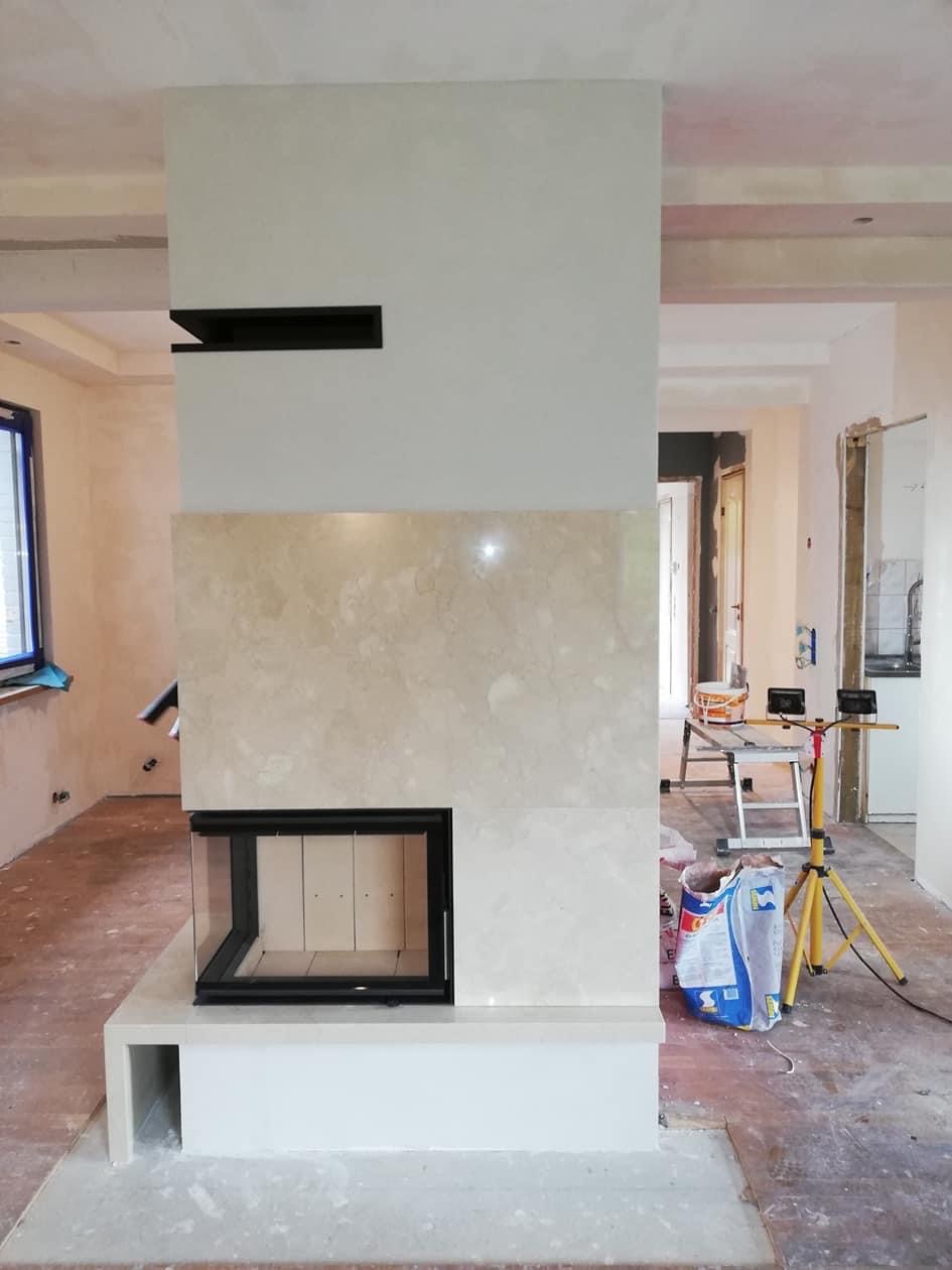 Wnętrze w trakcie wykańczania z obudowanym wkładem kominkowym z beżowego marmuru, widoczny fragment czarnej półki nad obudową, podłoga częściowo wyłożona, reszta w trakcie prac, widoczne narzędzia...