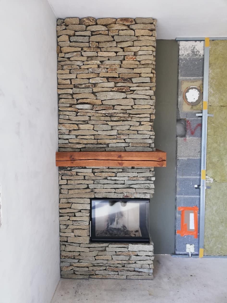 Wnętrze w trakcie wykończenia: obudowa z kamienia naturalnego wokół wkładu kominkowego z drewnianą półką, widoczne fragmenty ściany z ociepleniem.