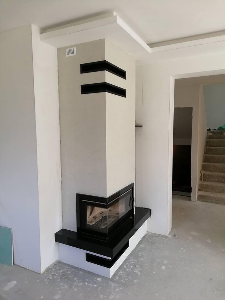 Nowoczesny narożny wkład kominkowy z czarnym obramowaniem i półkami w obudowie, w surowym wnętrzu z widocznymi schodami.