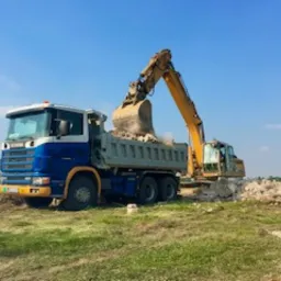 Żółta koparka załadowuje gruz na niebiesko-białą ciężarówkę Scania, w tle zielone pole i błękitne niebo.