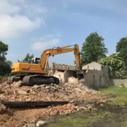 Żółta koparka Liebherr burzy fragment murowanego budynku z gruzem i pyłem w powietrzu, otoczona zielenią i błękitnym niebem.