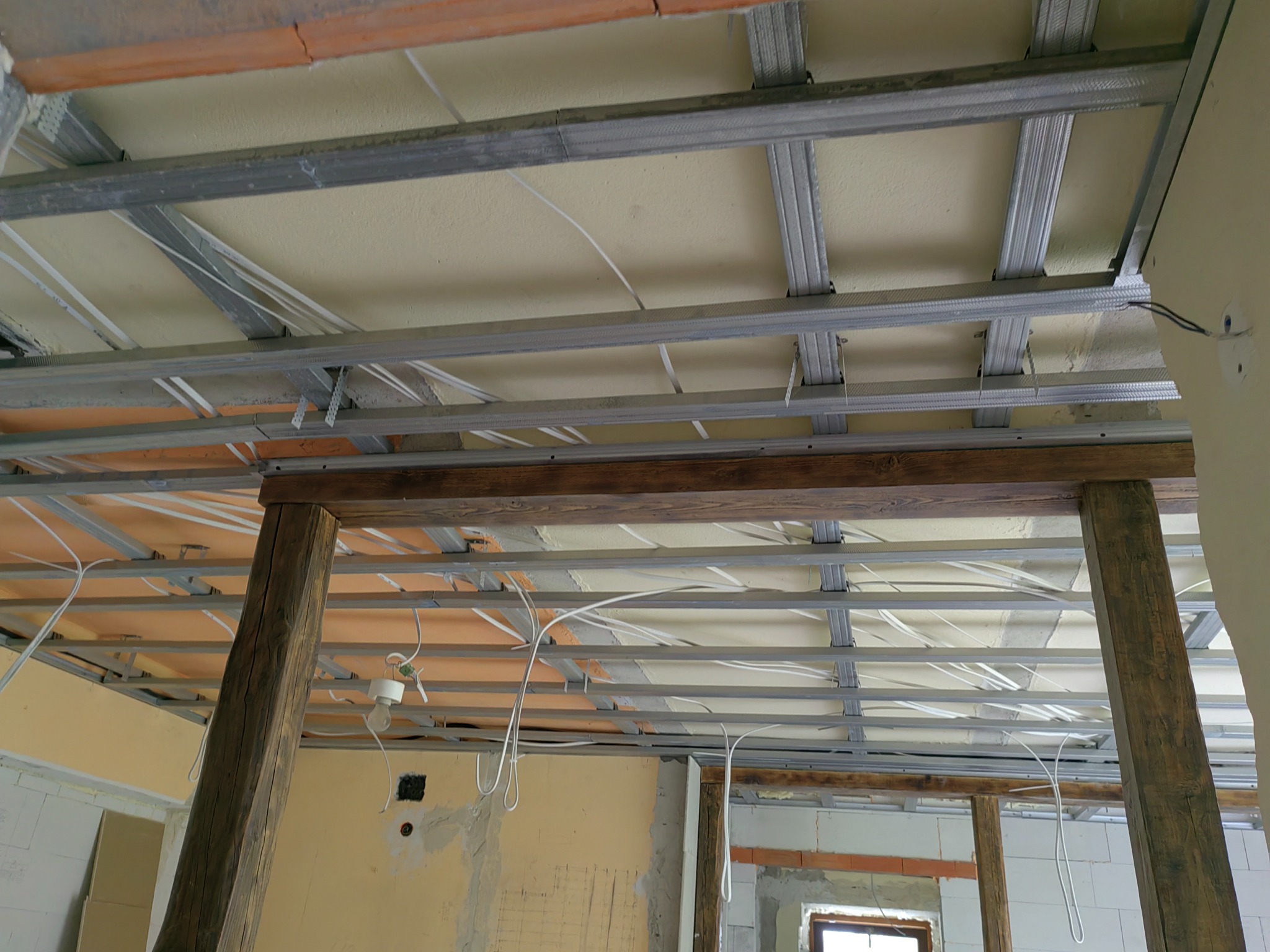 Montaż metalowej konstrukcji podwieszanego sufitu w pomieszczeniu z drewnianymi belkami, widoczne przewody elektryczne i niedokończone ściany.