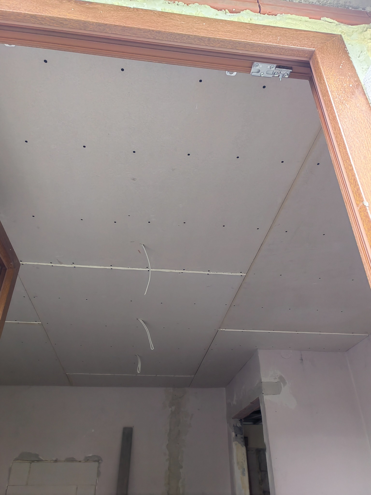 Montaż sufitu podwieszanego z płyt kartonowo-gipsowych, widoczne przewody elektryczne wystające z sufitu, drewniana rama drzwiowa.