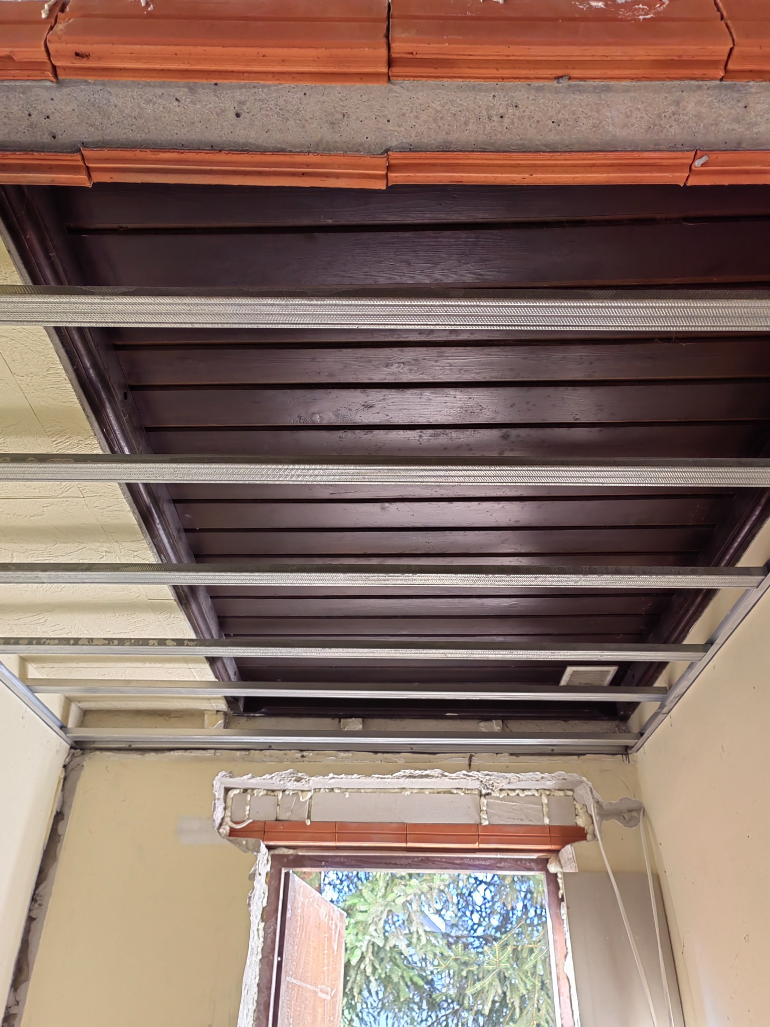 Montaż podwieszanego sufitu drewnianego z widoczną metalową konstrukcją nośną i oknem w tle, w trakcie prac wykończeniowych.