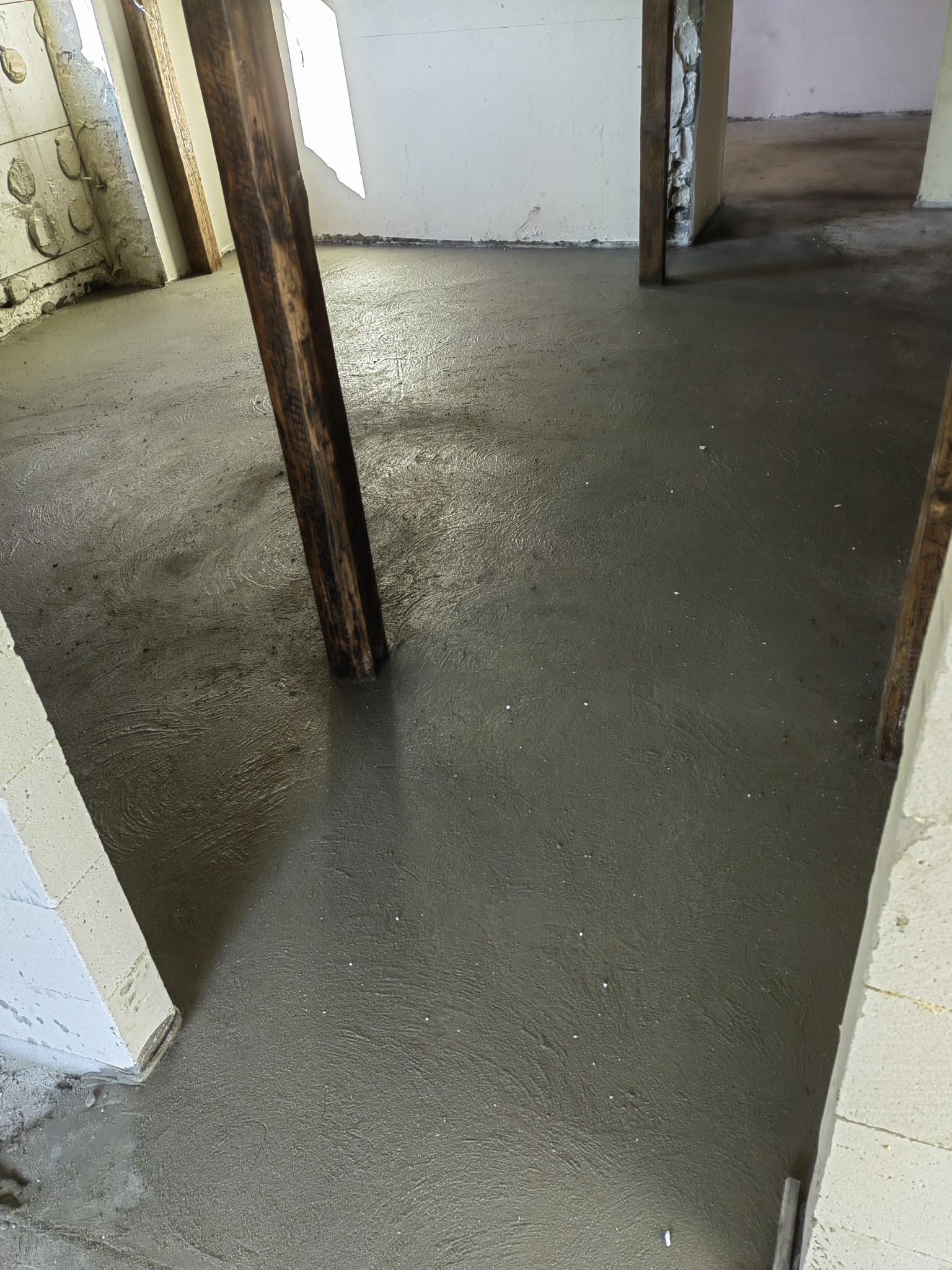 Świeżo wylana, mokra wylewka podłogowa w surowym wnętrzu z drewnianymi słupami i fragmentami ścian z betonu komórkowego.