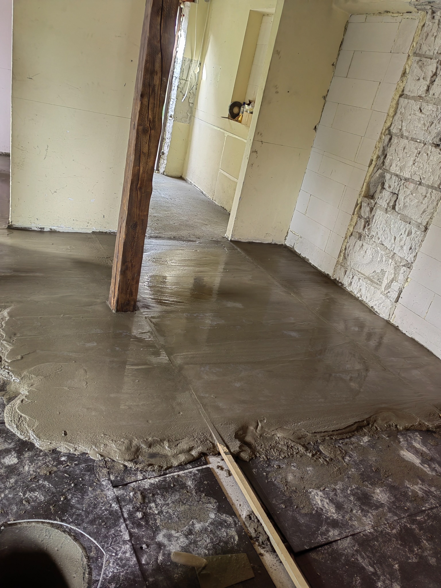 Wylewka betonowa na podłodze w trakcie wykonywania, widoczny drewniany filar wystający z betonu, ściany w trakcie wykończenia.