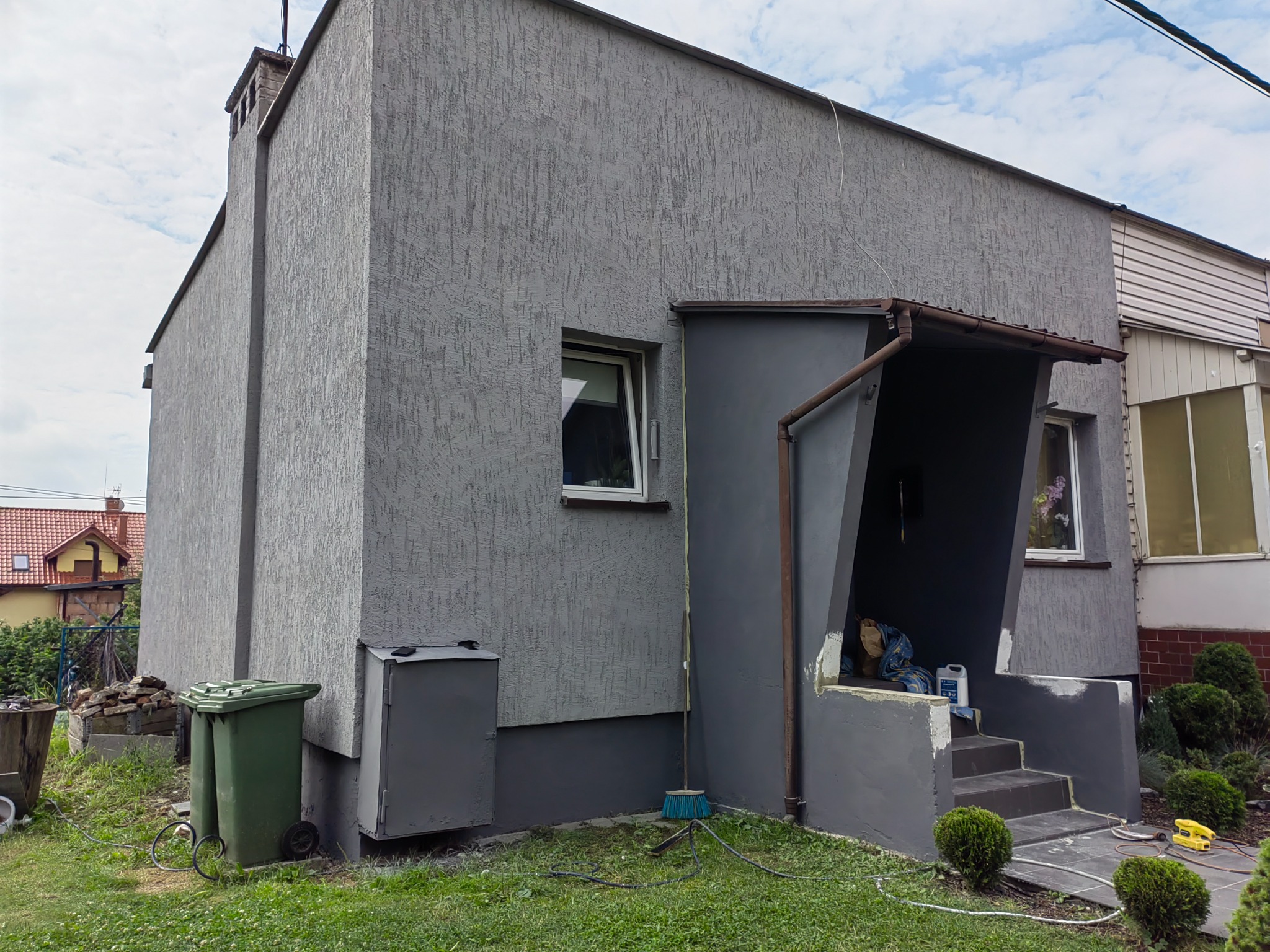 Szary dom z otynkowaną elewacją, widoczny z zewnątrz, z zielonymi pojemnikami na śmieci, miotłą i niedokończonymi pracami malarskimi przy wejściu.
