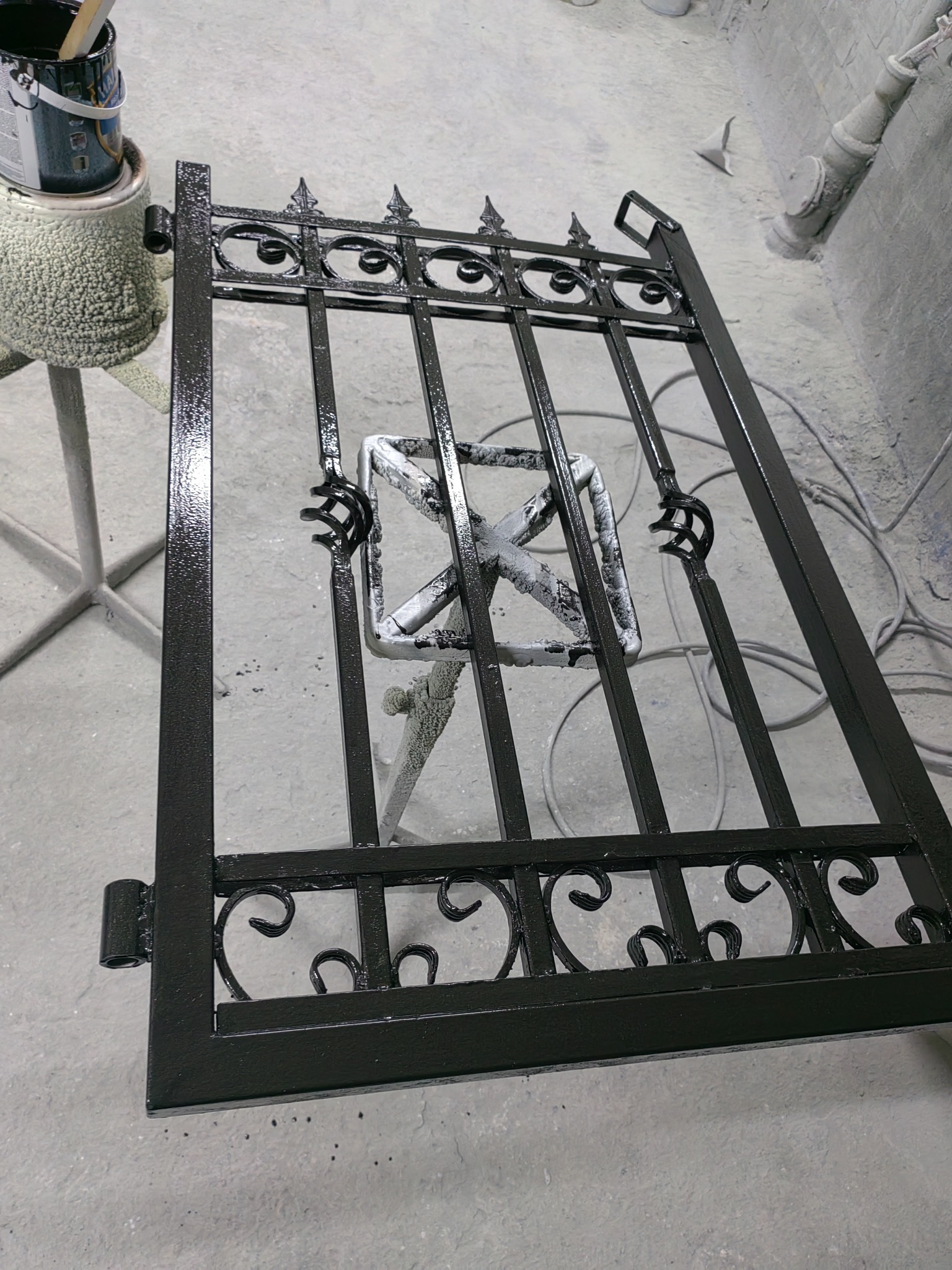 Świeżo pomalowana na czarno, ozdobna, metalowa furtka leży na stojaku w warsztacie, obok puszka z farbą i pędzel.