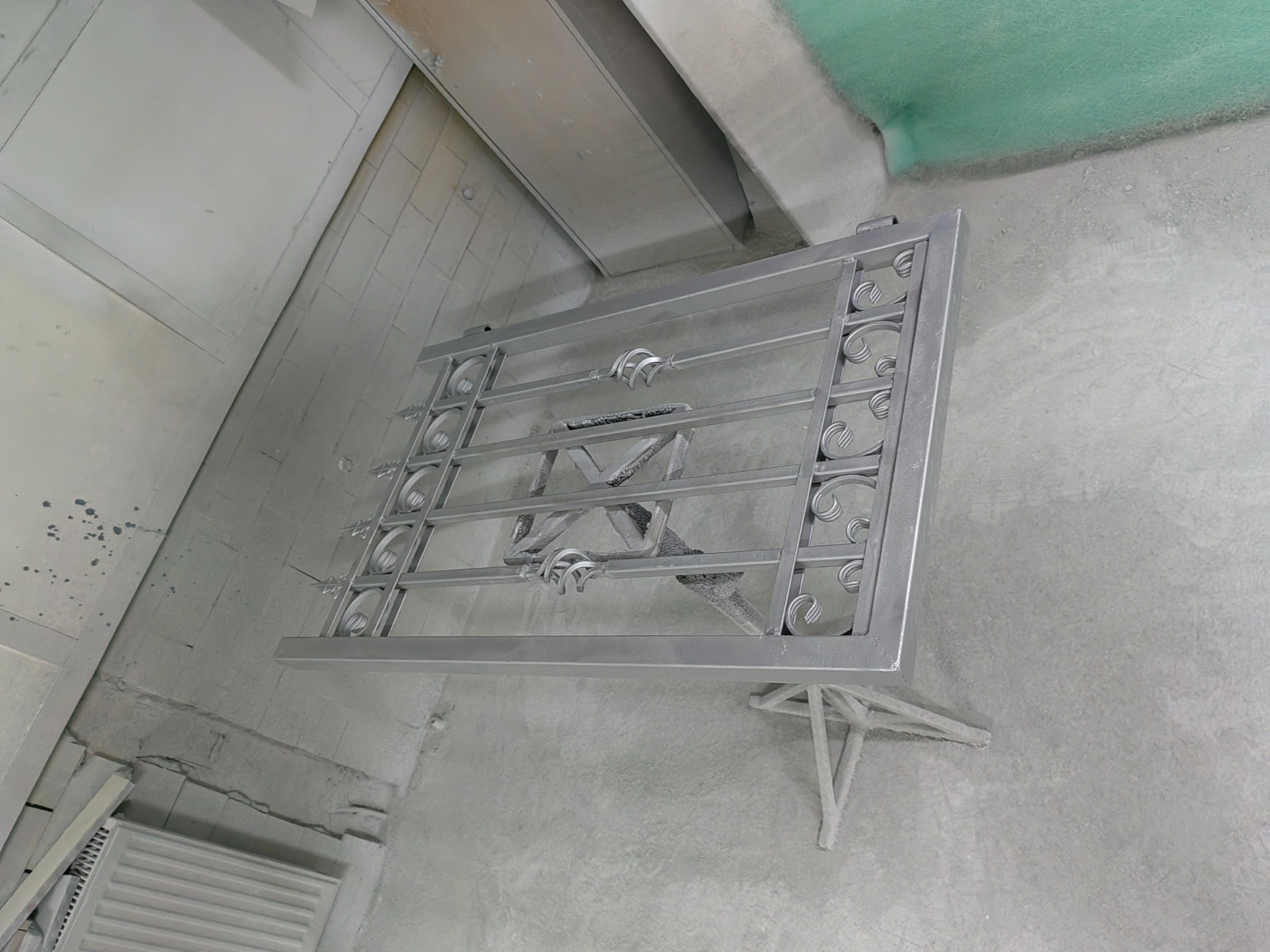 Metalowa, ozdobna krata okienna, świeżo pomalowana na srebrno, wisząca w warsztacie lub garażu z widocznymi śladami farby na ścianach i podłodze.