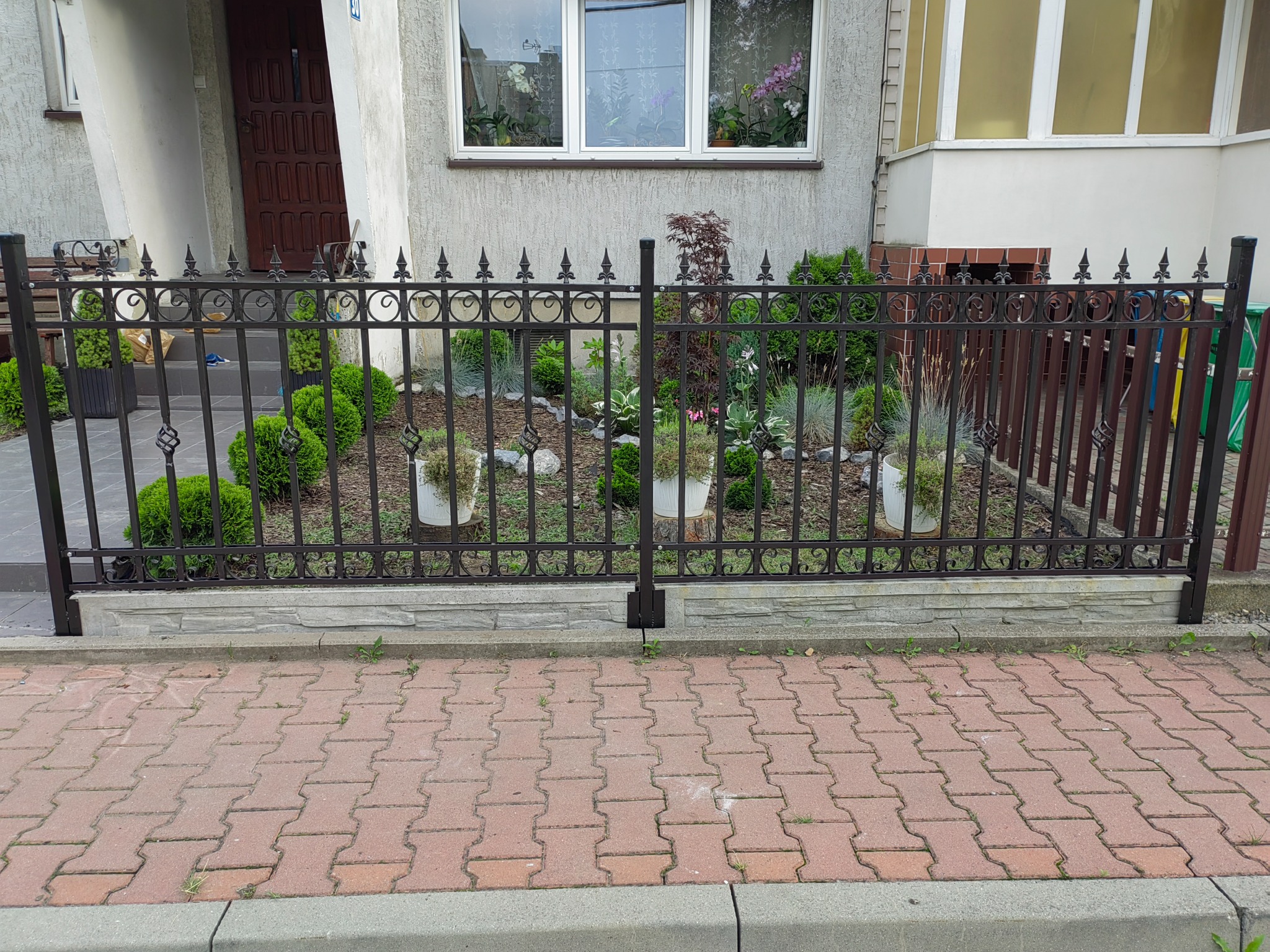 Kute, czarne ogrodzenie posesji z ozdobnymi detalami, oddzielające ogródek z roślinami od chodnika z czerwonej kostki brukowej.