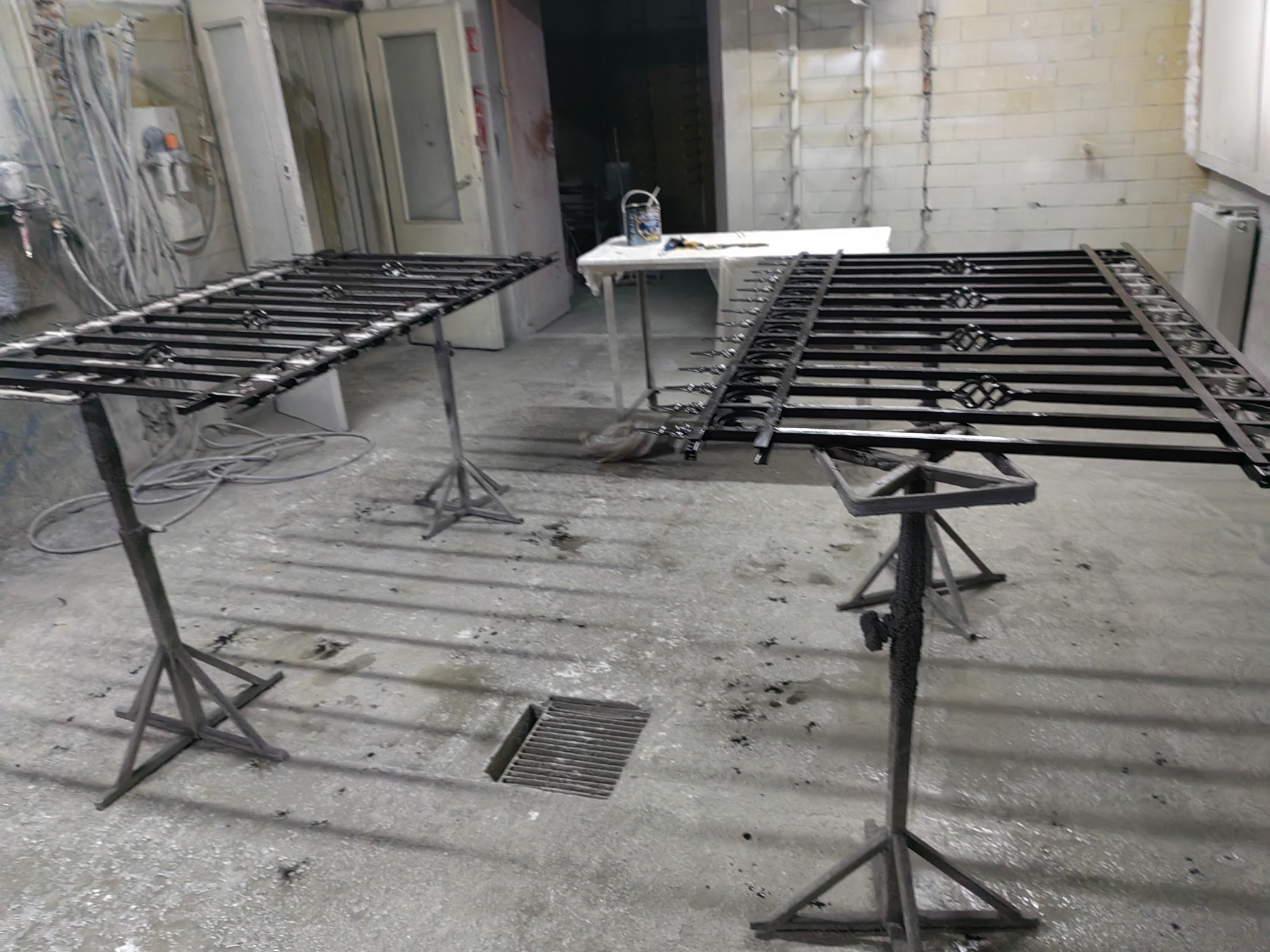 Dwie metalowe konstrukcje, prawdopodobnie fragmenty ogrodzenia lub balustrady, ustawione na metalowych stojakach w warsztacie, przygotowane do malowania lub po malowaniu, z widocznymi śladami farby...