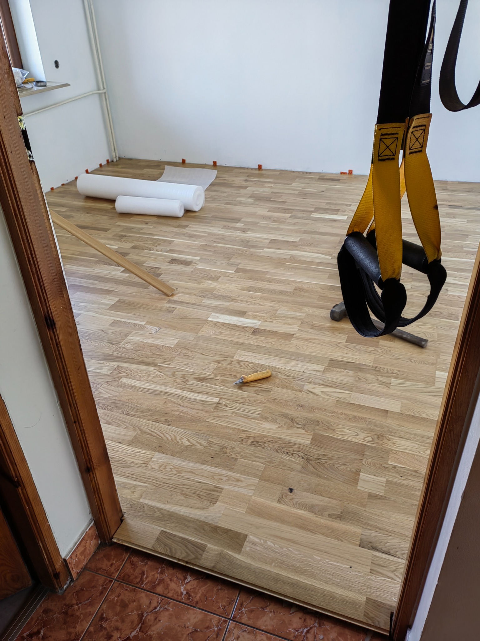 Parkiet ułożony w jodełkę, widoczne narzędzia: młotek, nóż do tapet, rolka podkładu, oraz pomarańczowe kliny dystansowe przy ścianach.