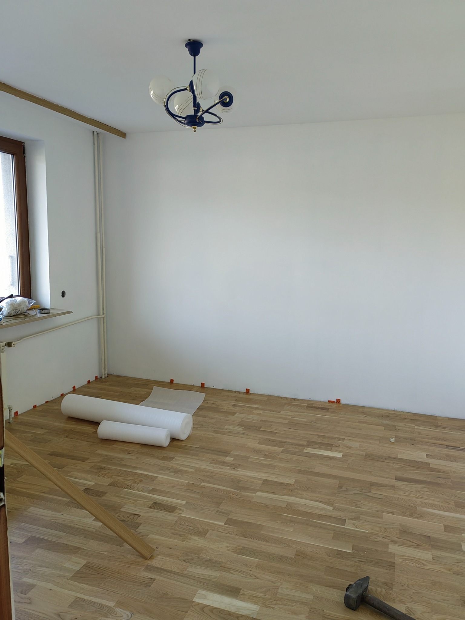 Ułożona drewniana podłoga w pustym pokoju, widoczne rolki materiału ochronnego, listwa przypodłogowa i młotek.