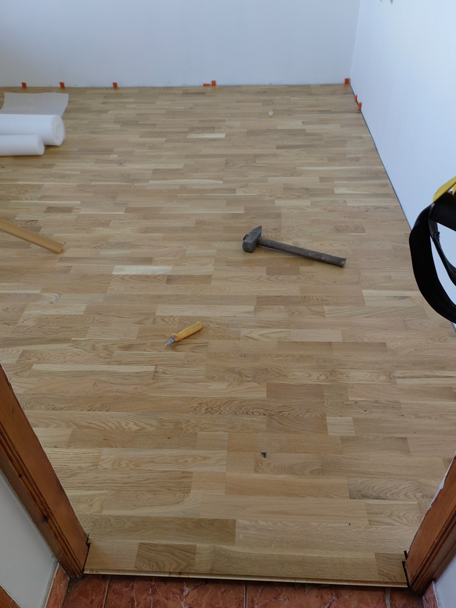 Ułożona drewniana podłoga z narzędziami: młotek i dłuto, widoczne rolki podkładu i pomarańczowe kliny dystansowe przy ścianie.