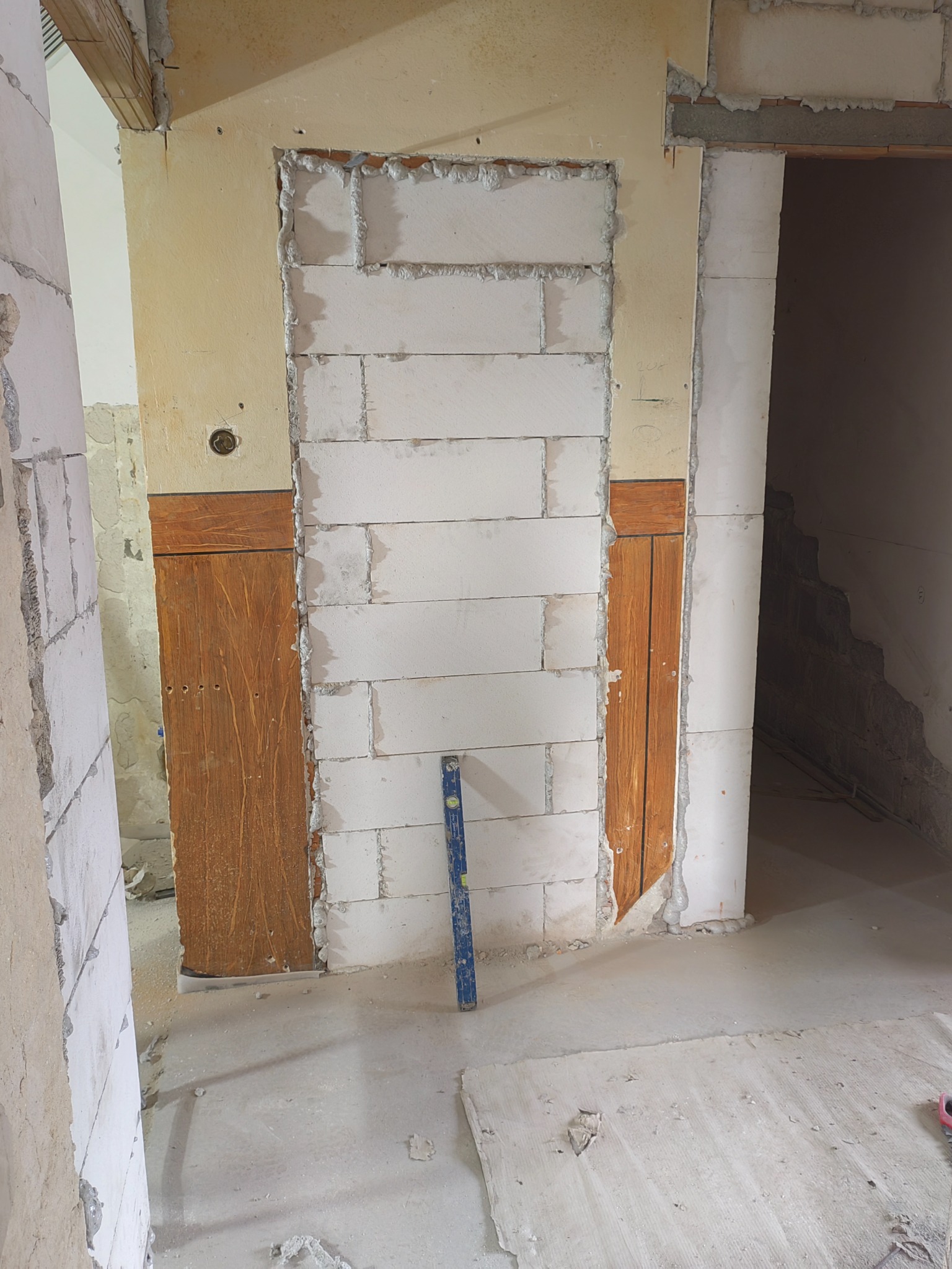 Wnętrze w trakcie remontu: murowane ściany z bloków, drewniane elementy wykończeniowe, poziomica oparta o ścianę, surowy beton na podłodze.