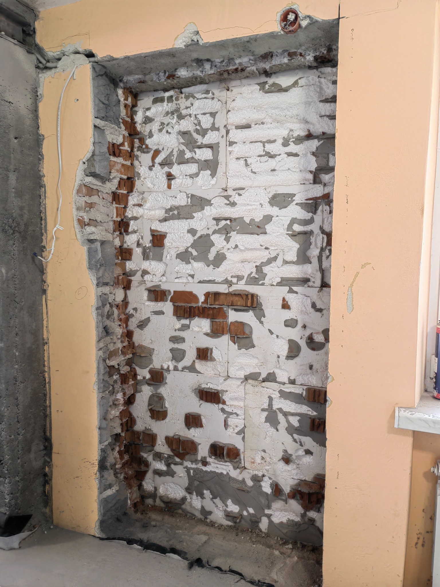 Otwór w ścianie z widoczną cegłą i resztkami tynku, przygotowany do dalszych prac wykończeniowych, z widoczną instalacją elektryczną w suficie.