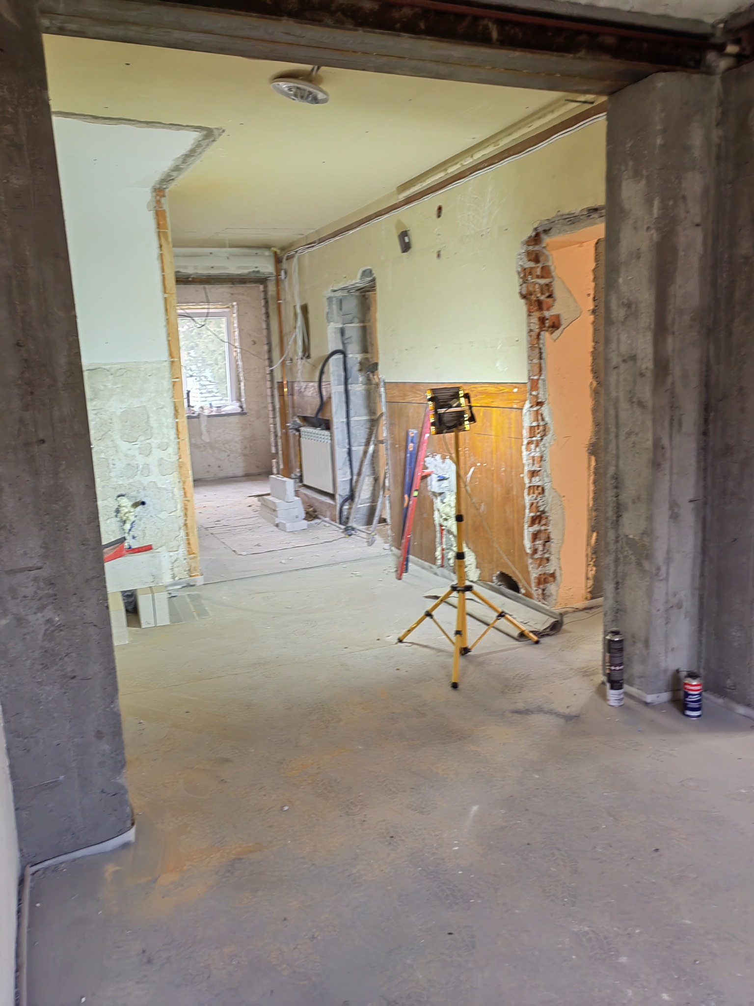 Wnętrze w trakcie remontu: odsłonięte cegły, surowy beton, żółty statyw oświetleniowy, częściowo otynkowane ściany i rozstawione narzędzia w pomieszczeniu z widocznymi śladami prac budowlanych.