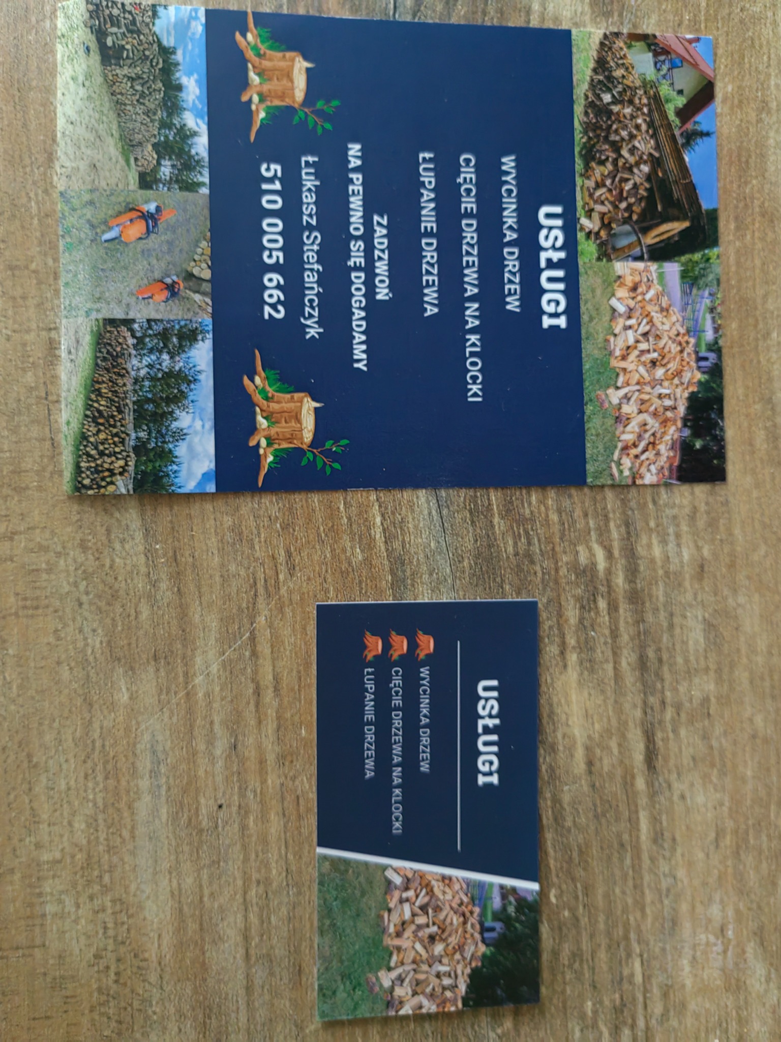 Dwie wizytówki firmy oferującej usługi związane z obróbką drewna, leżące na drewnianym stole. Na wizytówkach widnieją hasła: 'Usługi', 'Wycinka drzew', 'Cięcie drzewa na klocki', 'Łupanie drzewa'...