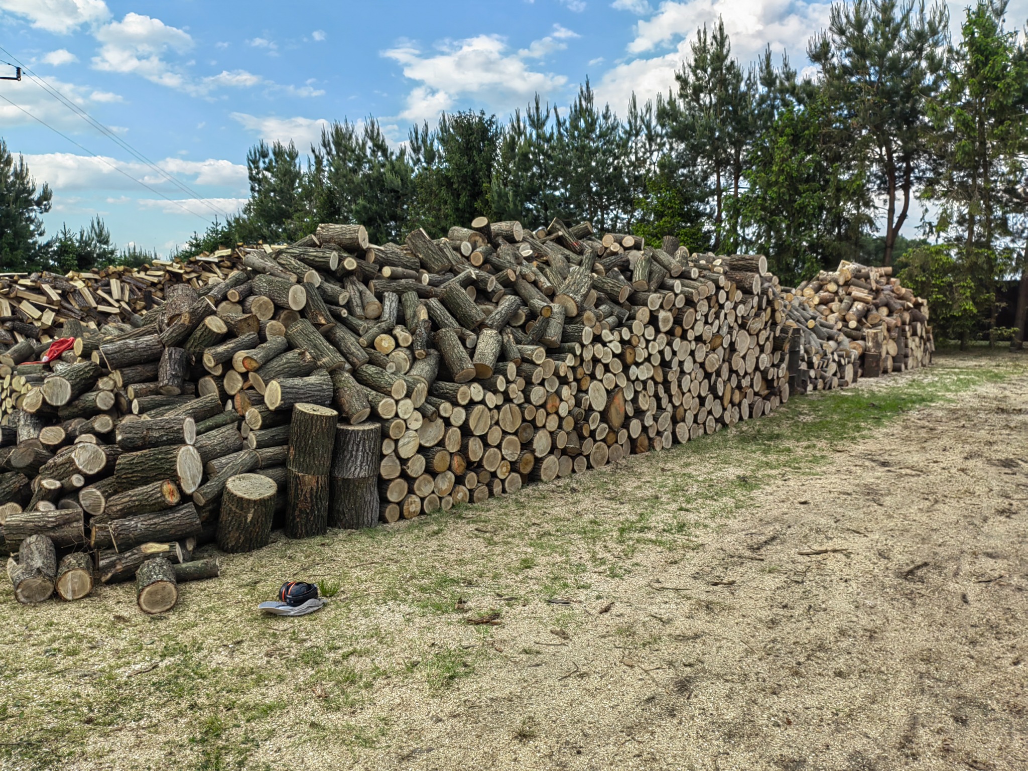 Stosy pociętego drewna ułożone na podwórku, w tle drzewa iglaste i błękitne niebo z chmurami, na pierwszym planie leżące na ziemi nauszniki ochronne.