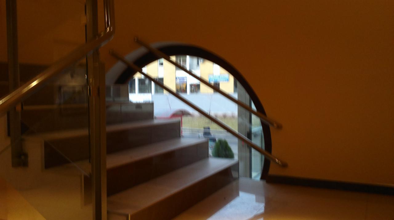 Schody z metalową poręczą i szklaną balustradą, widok z dołu na okno łukowe z widokiem na budynek.