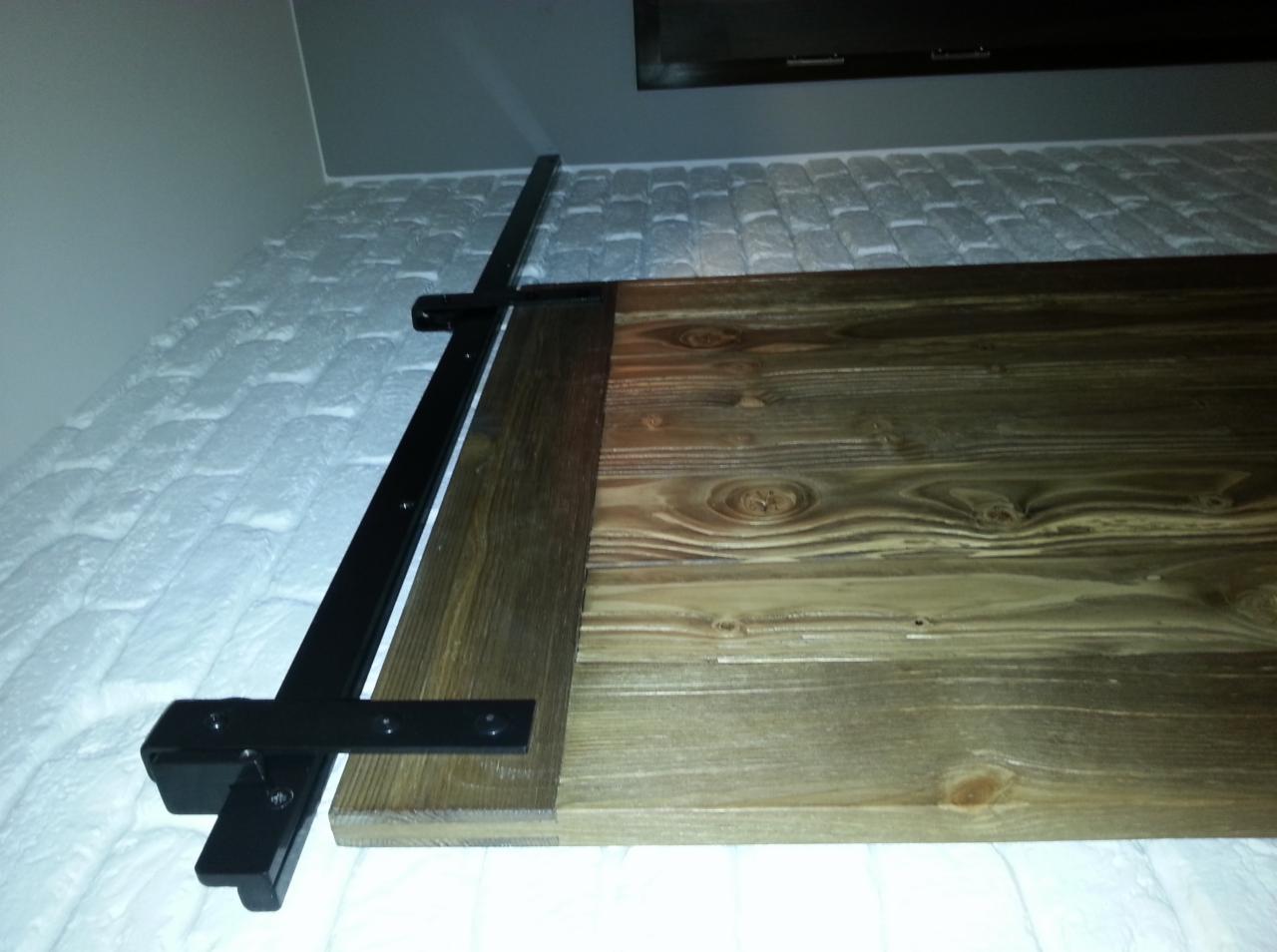 Fragment montażu drewnianej konstrukcji, prawdopodobnie balustrady, z czarnymi metalowymi elementami mocującymi na tle białej ściany z fakturą cegły.