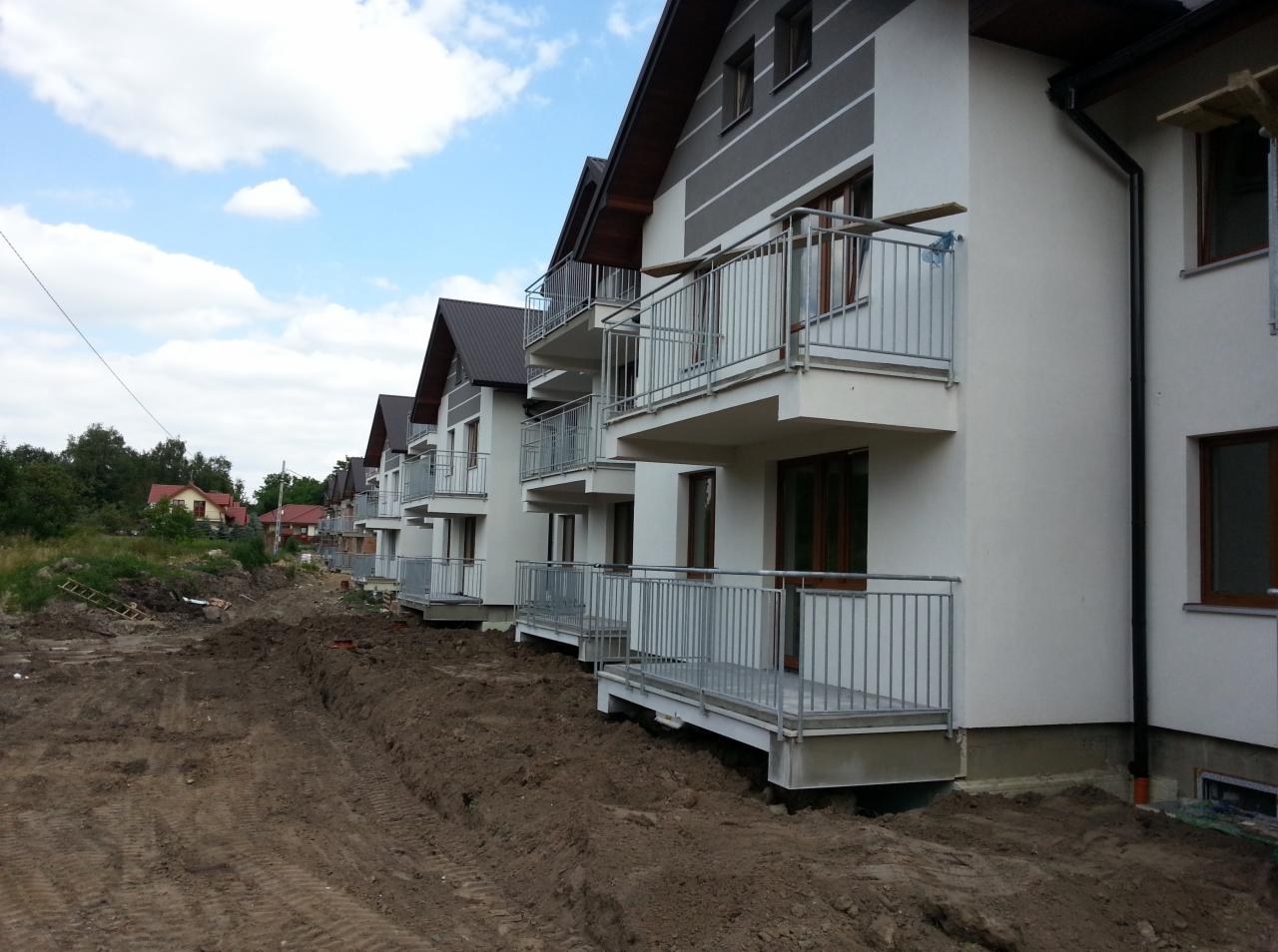 Rząd nowych domów szeregowych z balkonami z nierdzewnymi balustradami, widok na niezagospodarowany teren przed budynkami.
