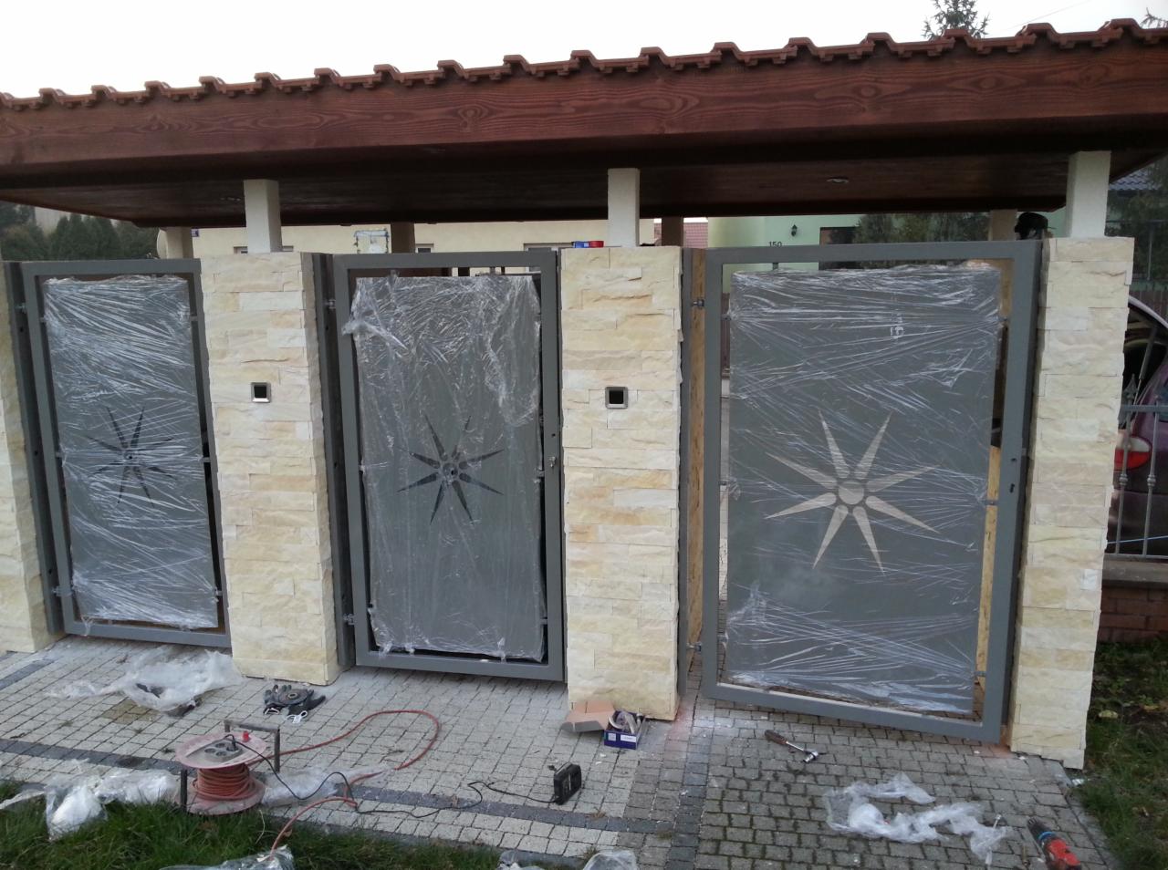 Trzy metalowe bramy z wyciętym motywem gwiazdy, osłonięte folią ochronną, zamontowane między kamiennymi słupkami, widoczne narzędzia montażowe i kostka brukowa.