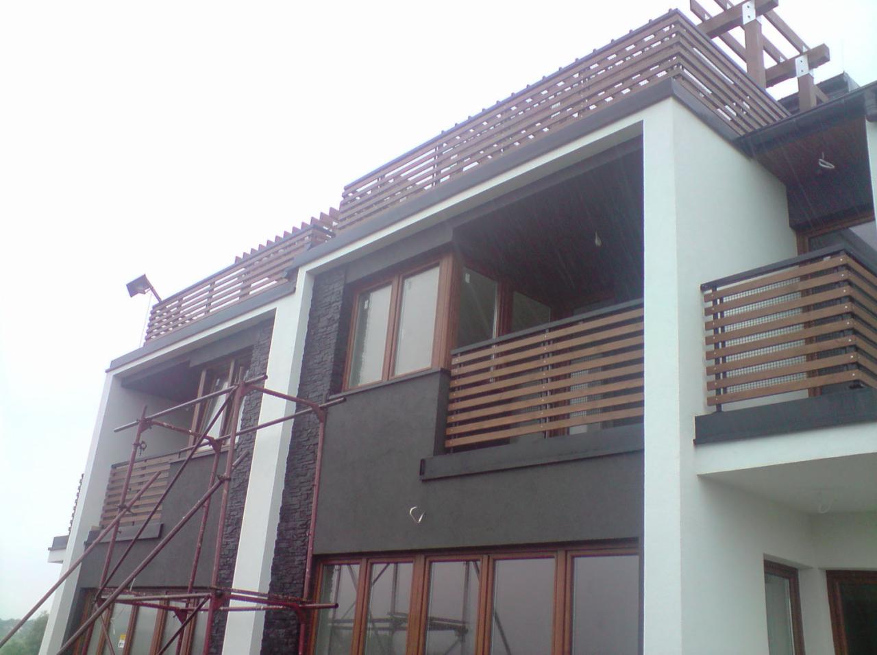 Nowoczesny dom z balkonami zabezpieczonymi poziomymi, drewnopodobnymi listwami, widoczne rusztowanie sugerujące trwające prace.