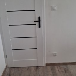 Firma Remontowo- Budowlana Edward Schulz - Białe drzwi z czarnymi akcentami i czarną klamką, zamontowane w jasnym wnętrzu z panelami podłogowymi. Widoczny włącznik i gniazdko elektryczne.