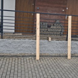 Firma Remontowo- Budowlana Edward Schulz - Wznoszenie murku oporowego z szarej kostki betonowej. Widoczne drewniane podpory, czarna balustrada i ułożona kostka brukowa. Etap budowy przed domem.