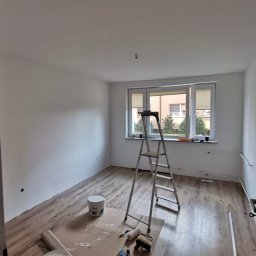 Firma Remontowo- Budowlana Edward Schulz - Pokój po malowaniu ścian na biało, z drabiną, farbami i narzędziami malarskimi na podłodze z paneli. Widok na okno z zewnątrz.