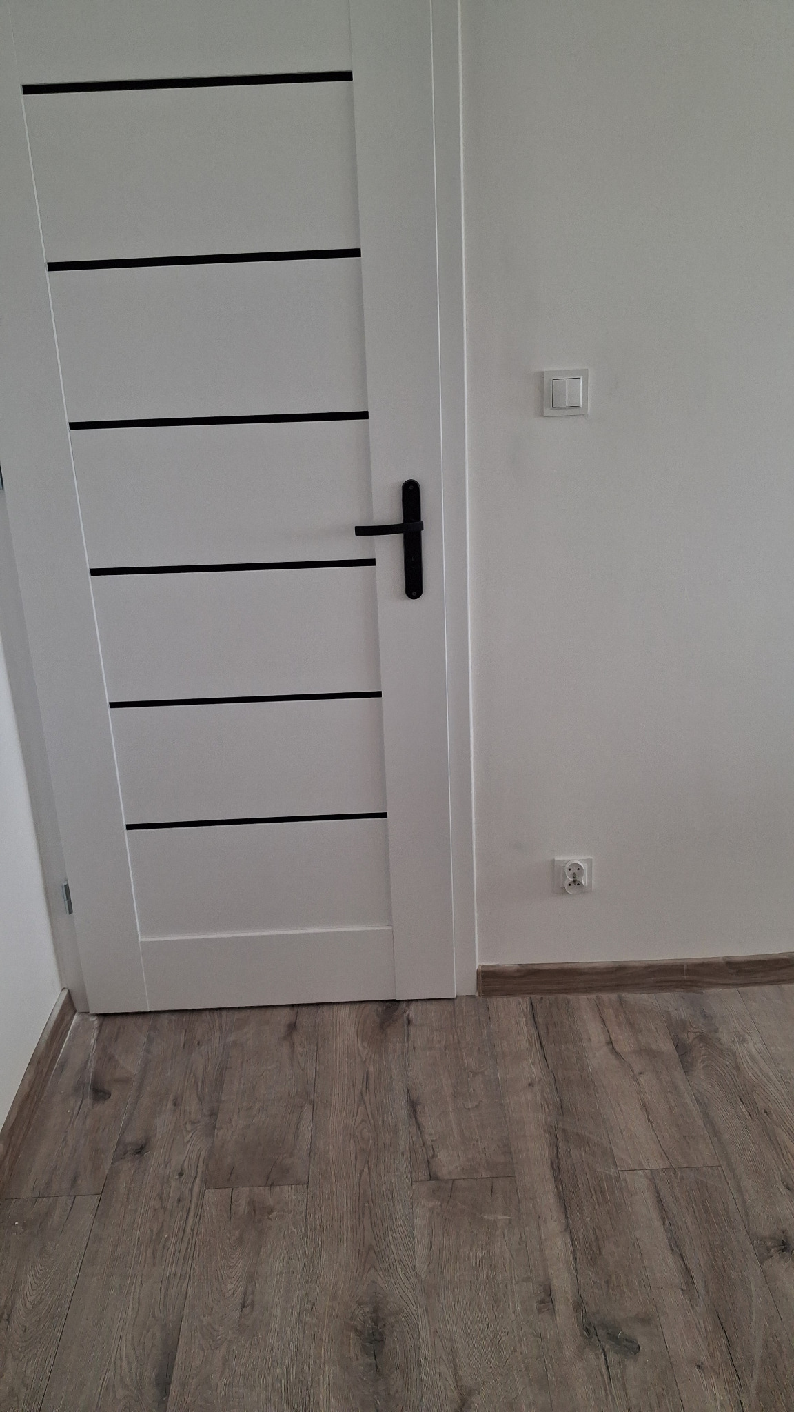 Białe drzwi z czarnymi akcentami i czarną klamką, zamontowane w jasnym wnętrzu z panelami podłogowymi. Widoczny włącznik i gniazdko elektryczne.