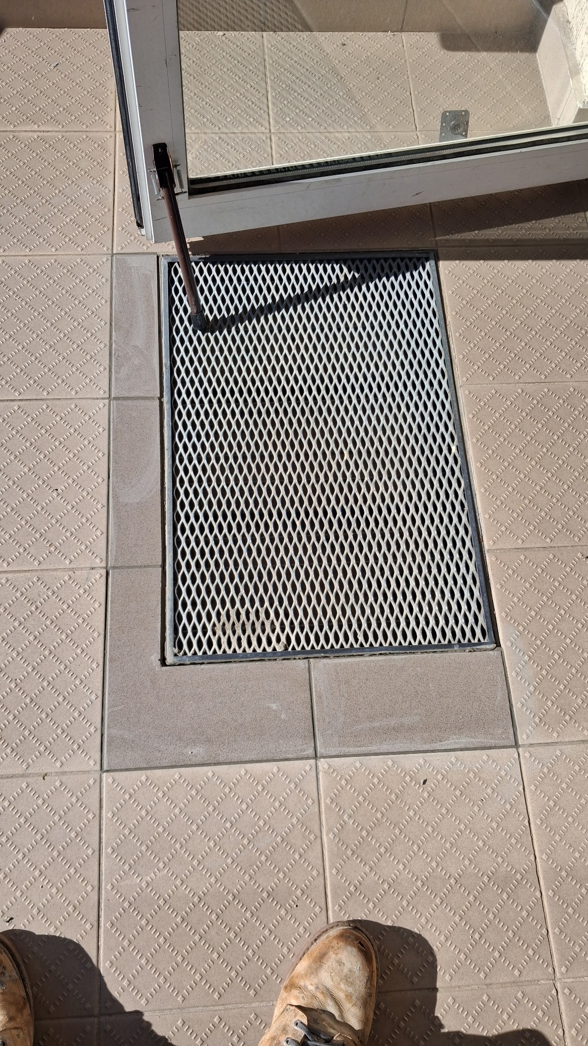 Kratka ściekowa wkomponowana w beżową, wzorzystą terakotę przy progu drzwi balkonowych. Widoczny fragment buta. Ujęcie z góry.